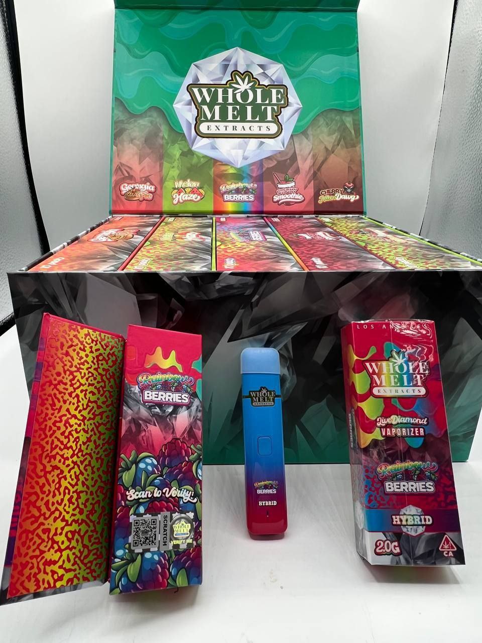 A display with multiple packaged cannabis products from the brand "Whole Melt Extracts." The image shows colorful branding and variety, including a vaporizer device and various boxes labeled "Import placeholder for 25." A QR code and hybrid product label are visible.