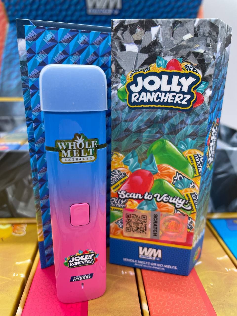 A blue and pink disposable vape pen labeled "Import placeholder for 26" and "Whole Melt Extracts" stands upright next to a box with colorful candy illustrations and the "Import placeholder for 26" logo. The box features a QR code and the text "Scan to Verify.