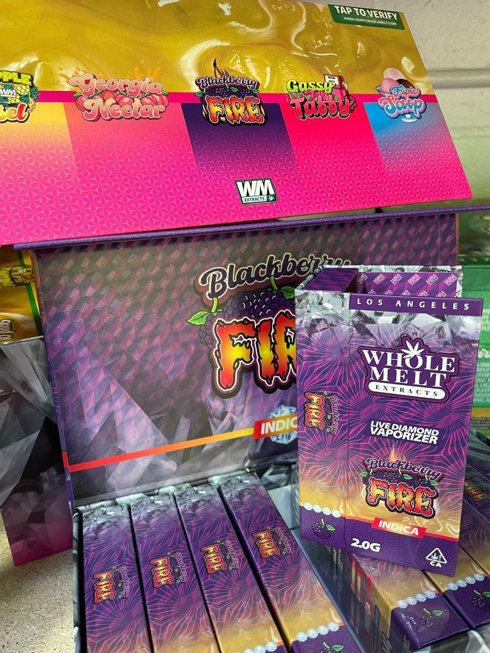 Photo of a display for a cannabis product called "Import placeholder for 50," by Whole Melt Extracts. The packaging is purple with a flame design. The display includes various strains and a box indicating it’s a live diamond vaporizer, indica, 2.0 grams.