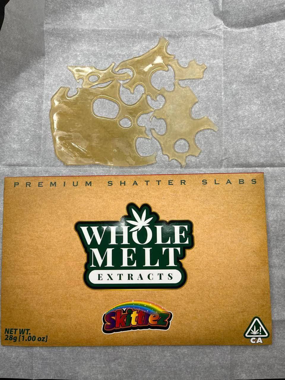 A package labeled "Whole Melt Extracts" and "Import placeholder for 59" with "Premium Shatter Slabs" is displayed beneath a sheet of light amber-colored, brittle cannabis concentrate. The package shows a net weight of 28g (1.00 oz) and has a cannabis warning symbol on the bottom right corner.